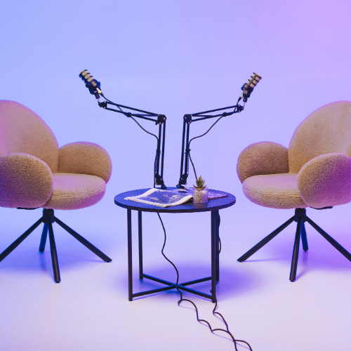 Two beige chairs face each other across a circular black table with two podcast microphones in a softly lit recording studio.